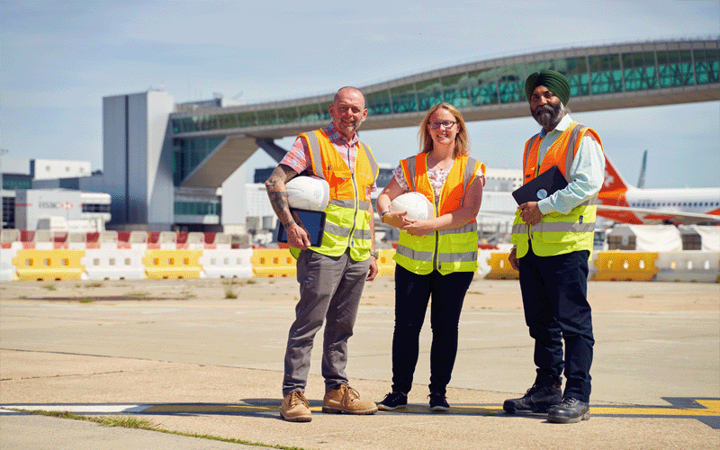Photo of three construction colleagues standing on the airfield with the pier 6 bridge behind them